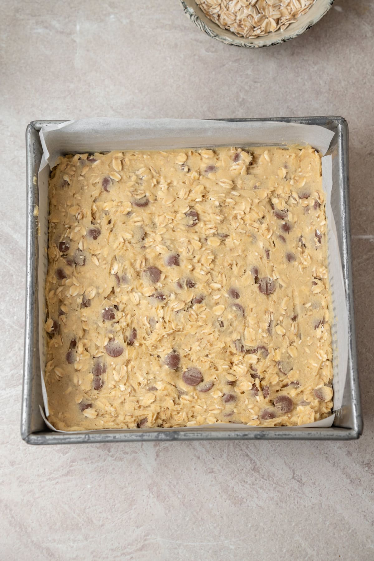 Oatmeal chocolate chip cookie dough pressed into a parchment-lined square baking pan to make bars.