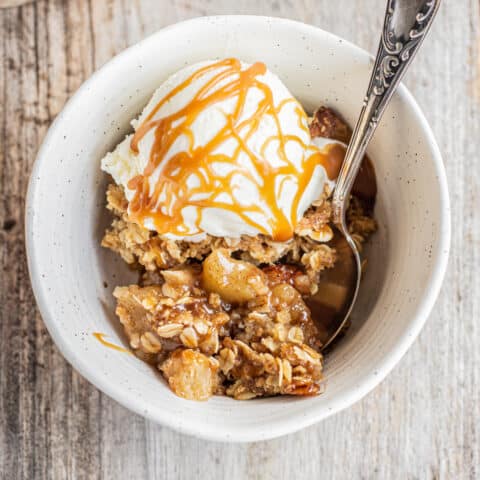 A bowl of warm salted caramel apple crisp topped with a scoop of vanilla ice cream and drizzled with caramel sauce. A spoon is placed in the bowl, and the dessert rests on a rustic wooden surface.