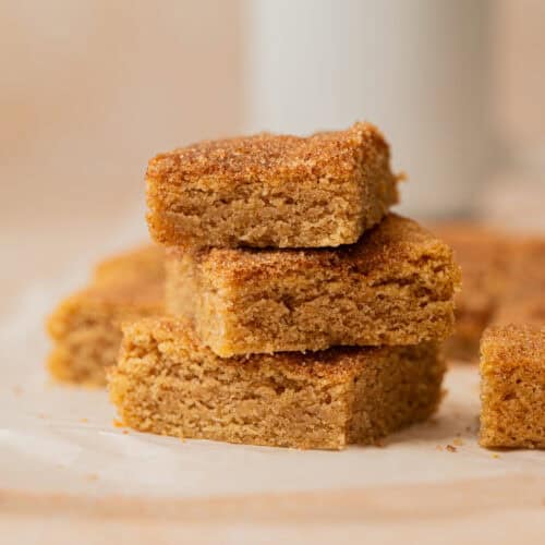Three stacked golden-brown snickerdoodle cookie bars with a crumbly texture, sitting on a piece of parchment paper. More bars and a blurred white mug are visible in the background.