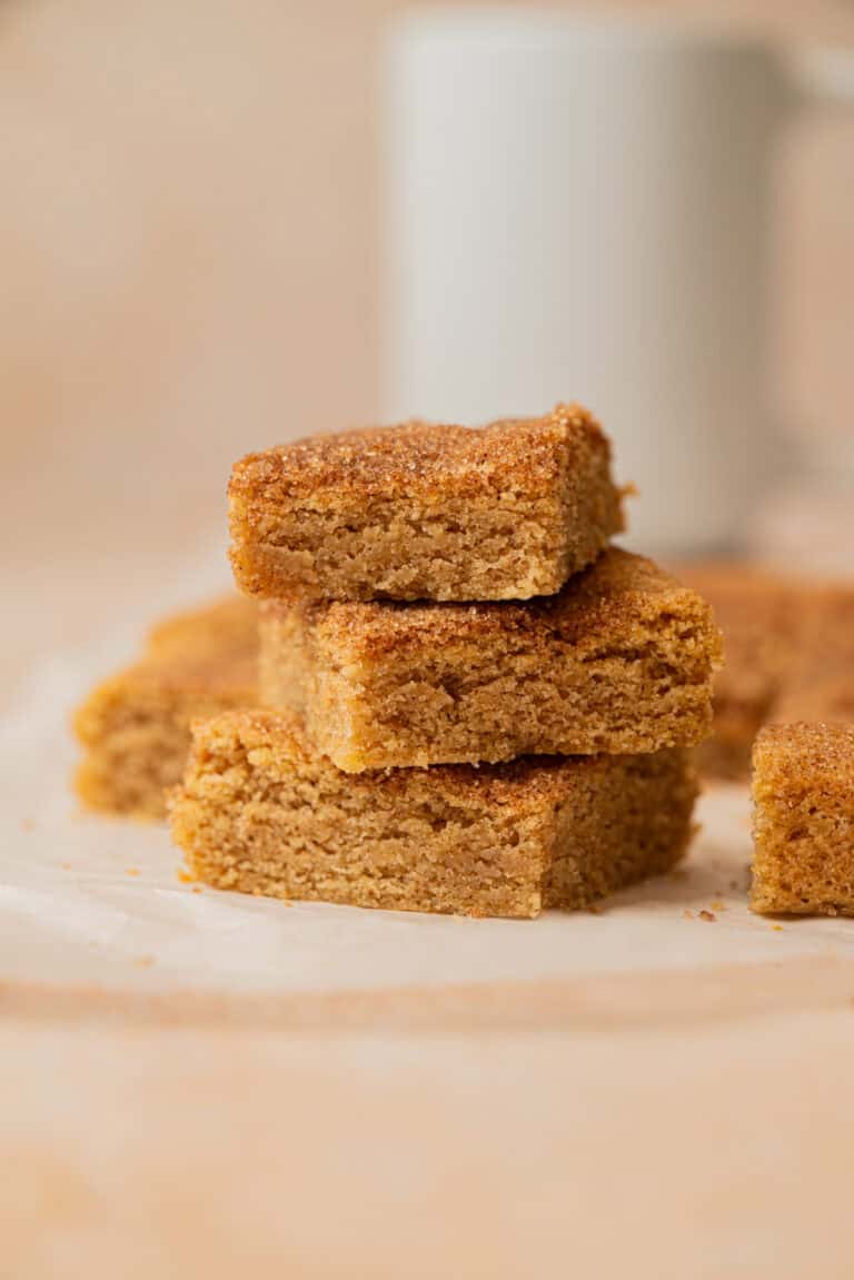 Three stacked golden-brown snickerdoodle cookie bars with a crumbly texture, sitting on a piece of parchment paper. More bars and a blurred white mug are visible in the background.