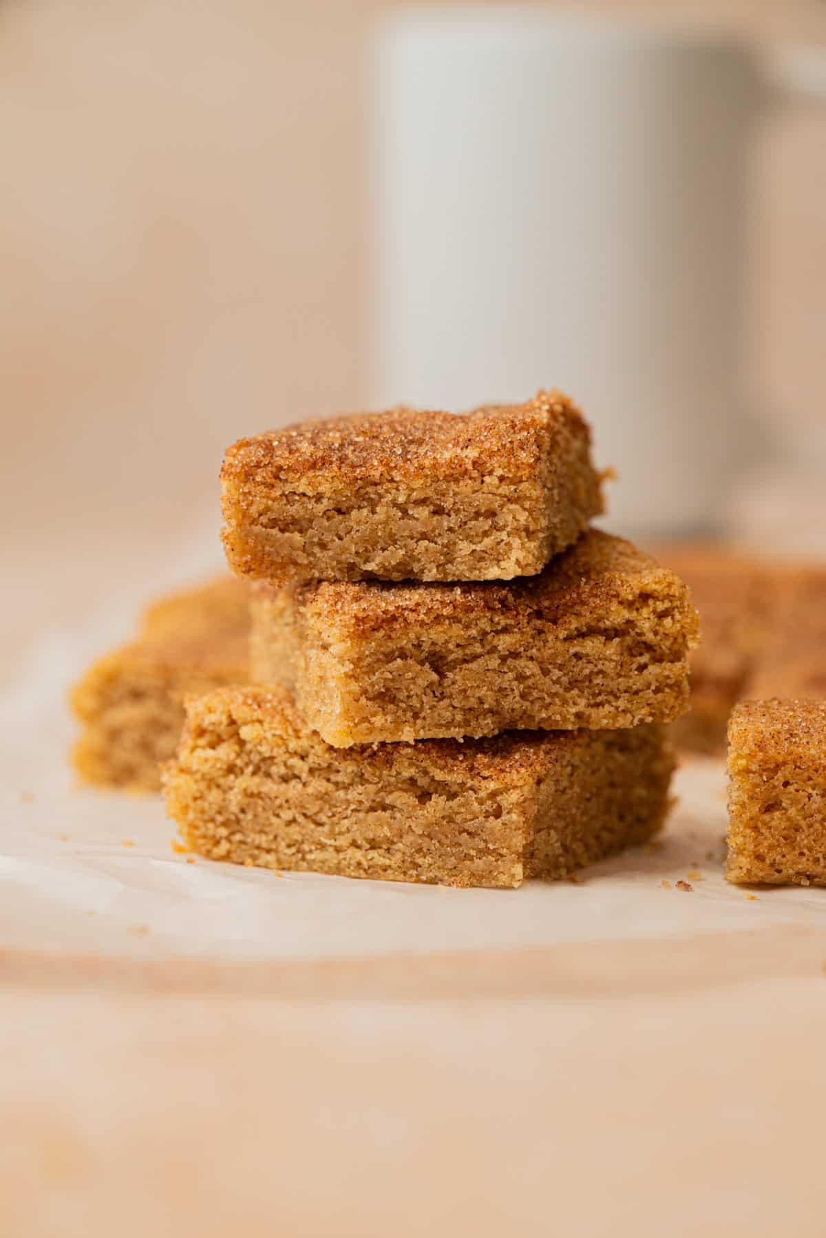 Three stacked golden-brown snickerdoodle cookie bars with a crumbly texture, sitting on a piece of parchment paper. More bars and a blurred white mug are visible in the background.