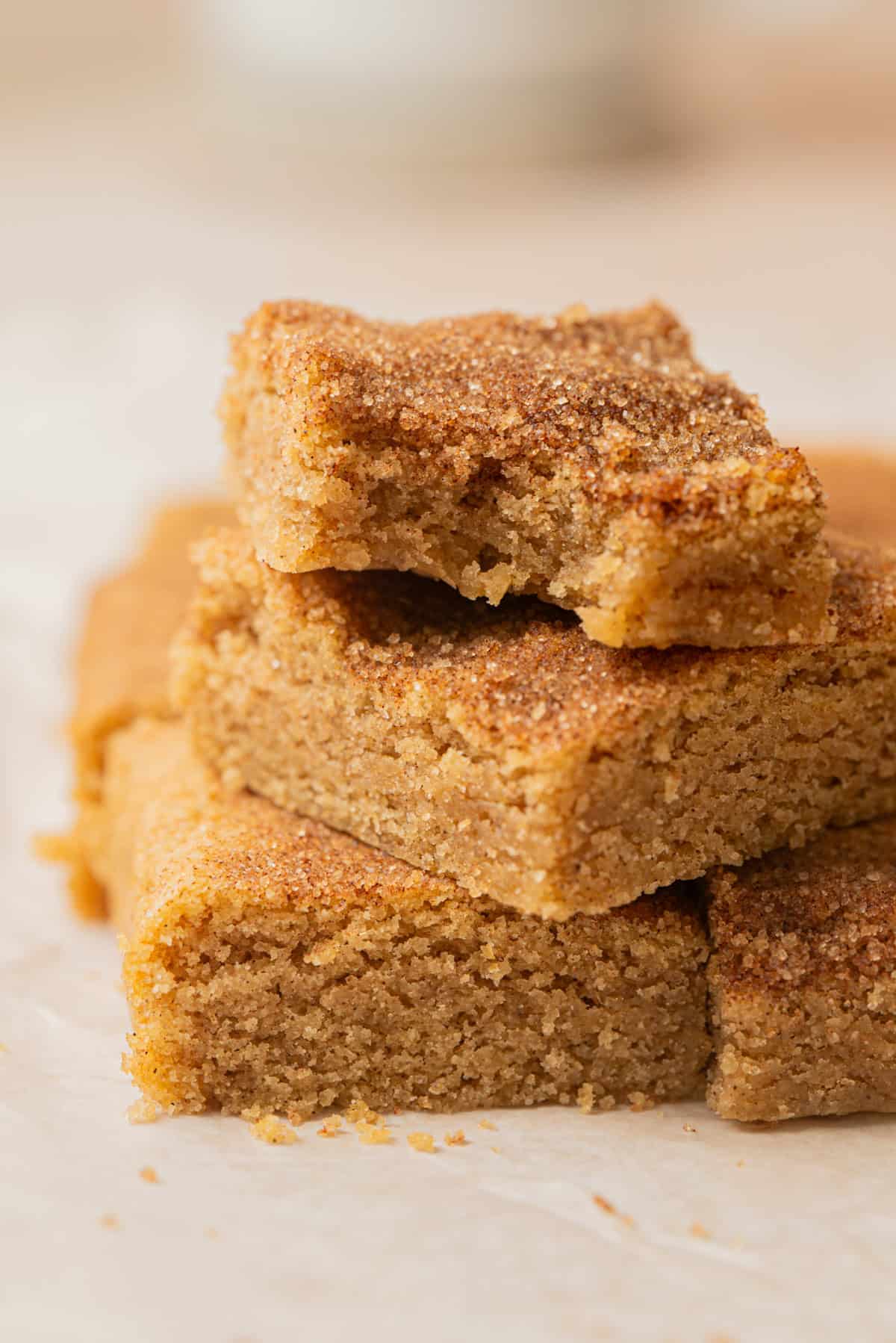 A close-up of a stack of golden brown, soft snickerdoodle cookie bars, with one bar on top showing a bite taken out, revealing a moist, crumbly texture. The bars rest on a piece of parchment paper.