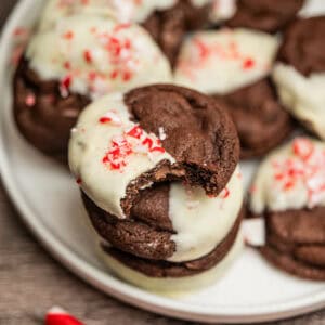A stack of peppermint chocolate cookies, partially dipped in white chocolate and sprinkled with crushed peppermint candy, with one cookie showing a bite taken out of it, on a white plate.