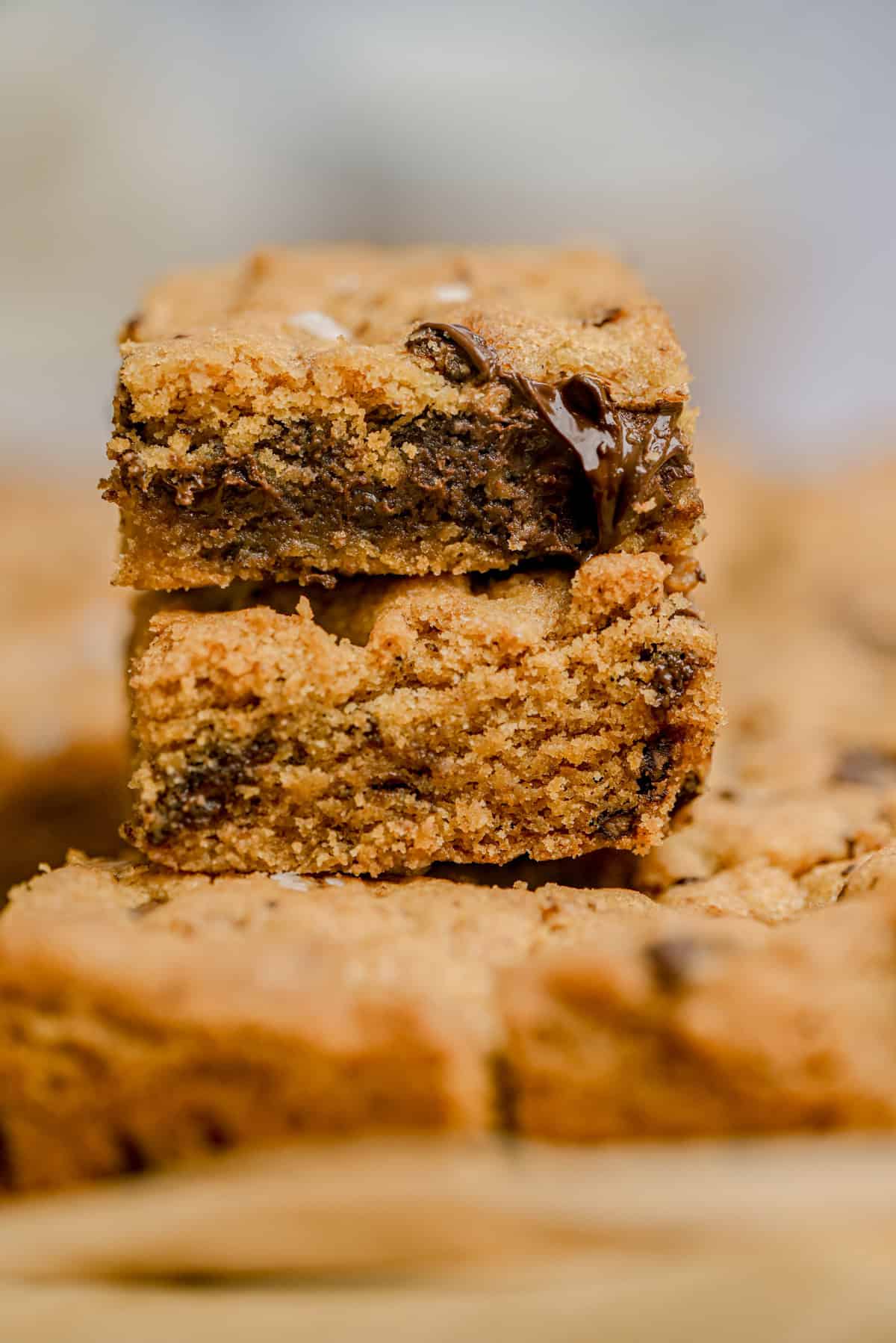 Two thick, chewy brown butter chocolate chip bars are stacked on top of each other, with melted chocolate visible in the top bar. More cookie bars are blurred in the background.