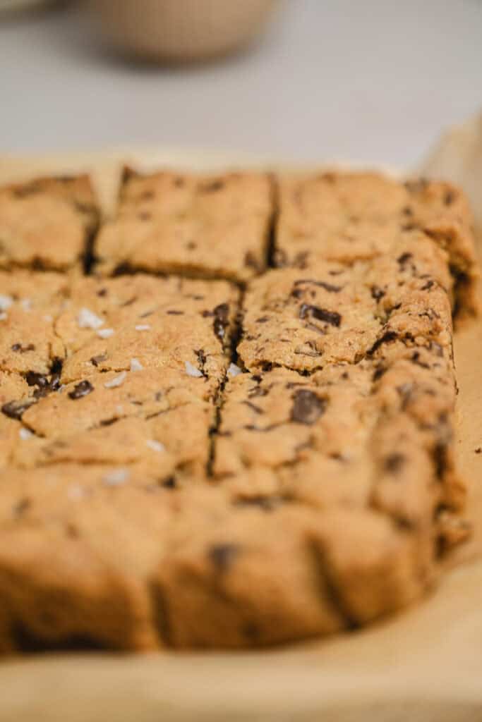 Close-up of freshly baked brown butter chocolate chip bars, cut into squares and resting on parchment paper, with a few flakes of sea salt sprinkled on top.