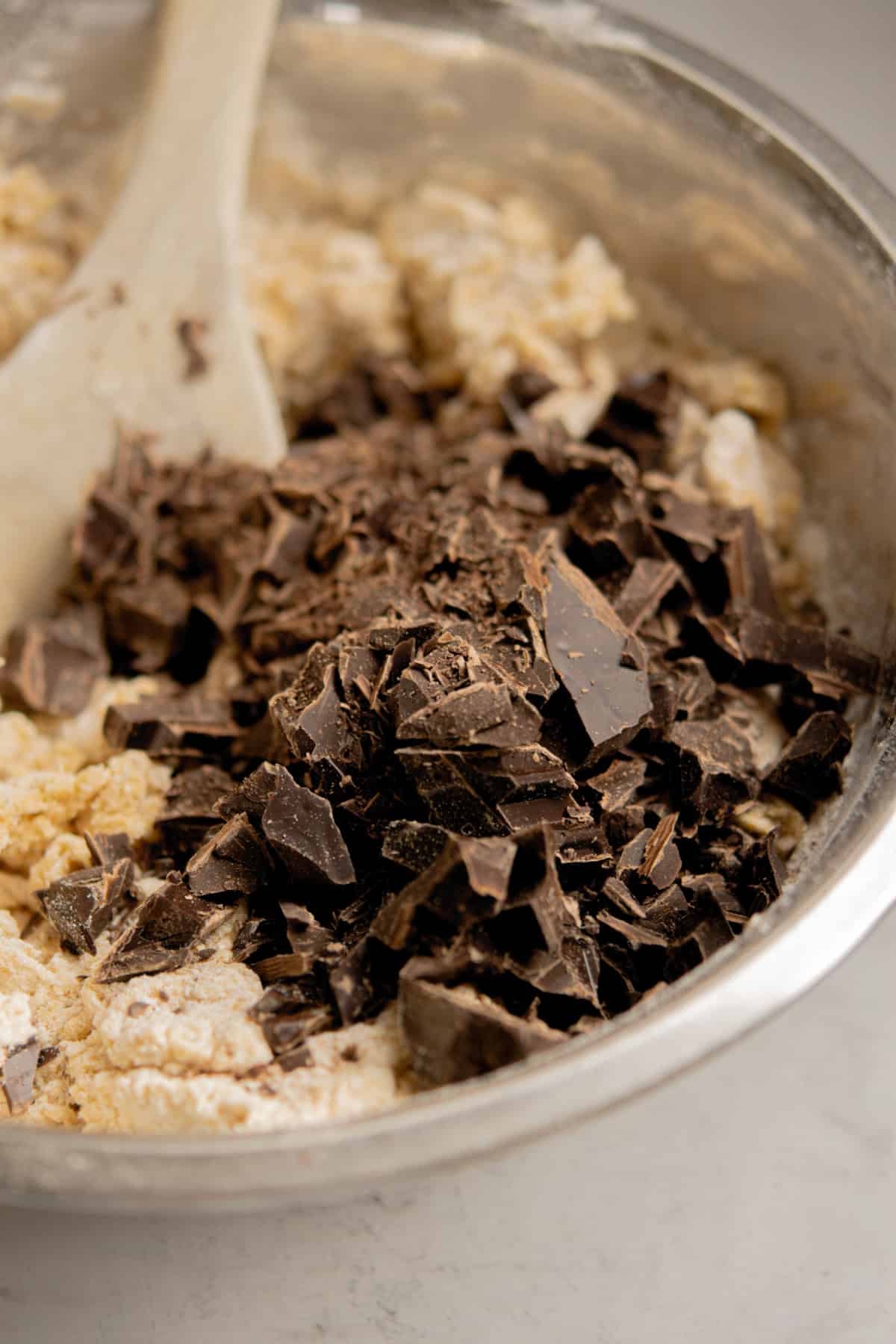 A metal mixing bowl filled with cookie dough for brown butter chocolate chip bars, topped with a pile of chopped dark chocolate pieces and a wooden spoon resting in the dough.