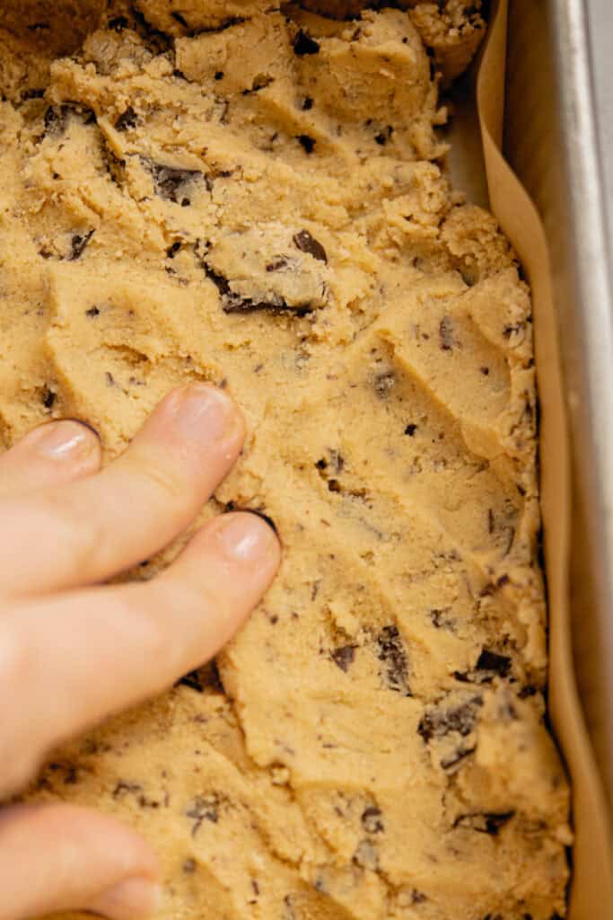 A close-up of a hand pressing brown butter chocolate chip bars dough into a parchment-lined baking pan.