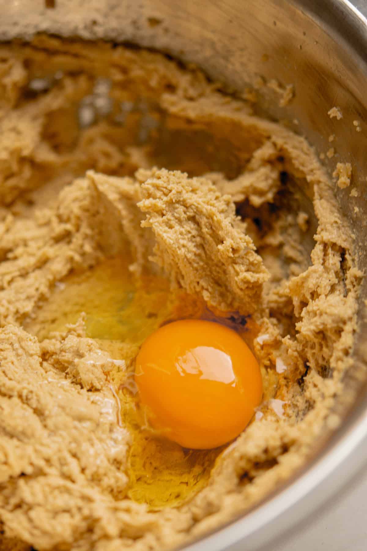 A close-up of cookie dough in a mixing bowl with a raw egg cracked on top, ready to be mixed in for brown butter chocolate chip bars.