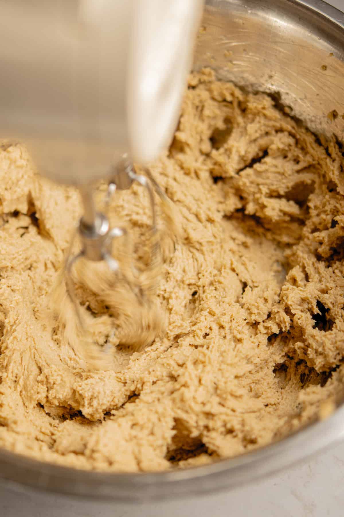 A close-up of an electric hand mixer blending cookie dough for brown butter chocolate chip bars in a large metal bowl. The dough appears partially mixed, with the mixer’s beaters in motion.