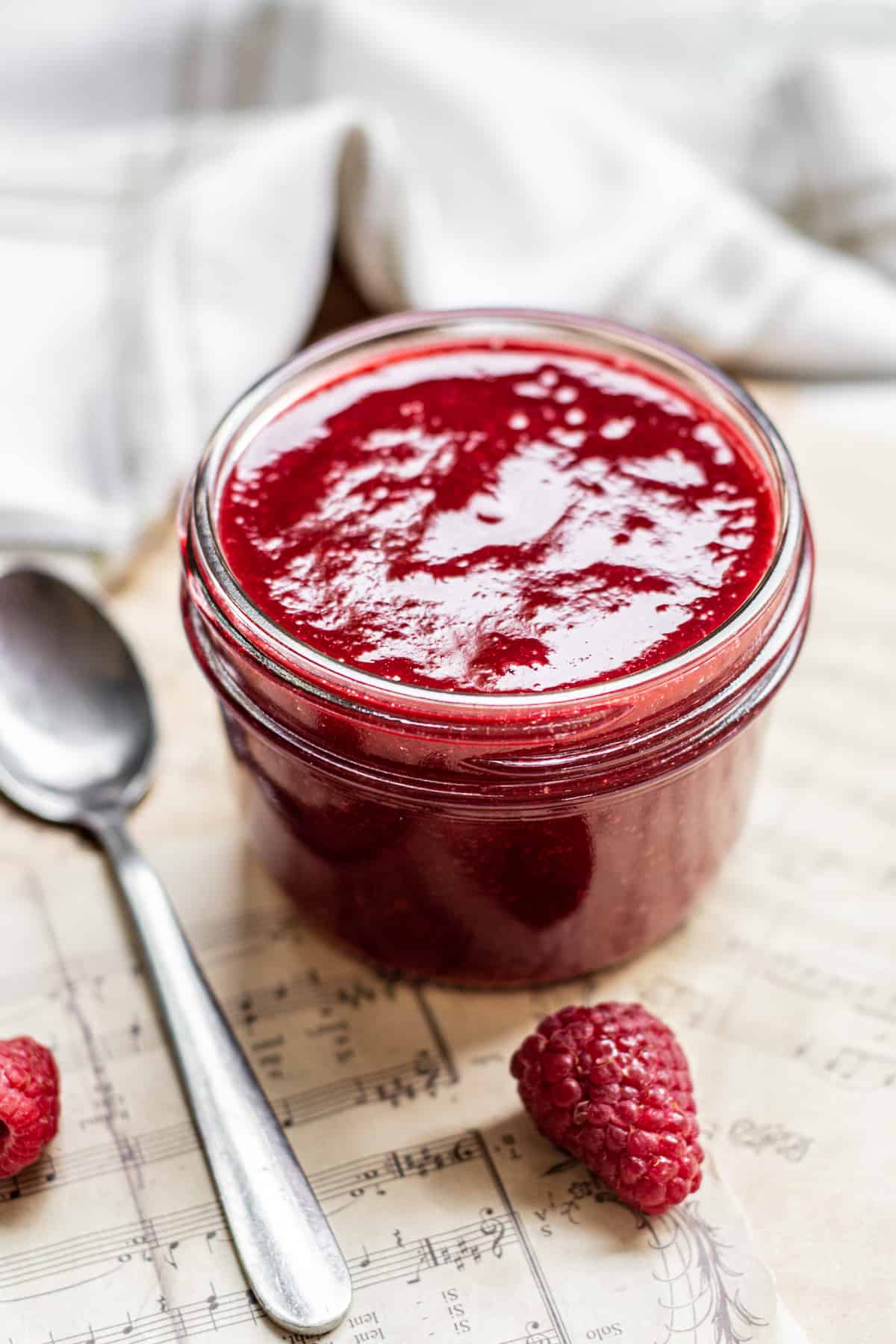 A glass jar filled with vibrant raspberry coulis sits on sheet music, next to a metal spoon and fresh raspberries, with a folded white cloth in the background.