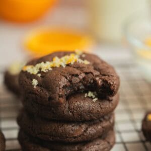 A stack of orange chocolate cookies sits on a cooling rack, with the top cookie partially bitten and sprinkled with sugar and orange zest. The background is softly blurred, showing hints of orange and a glass of milk.