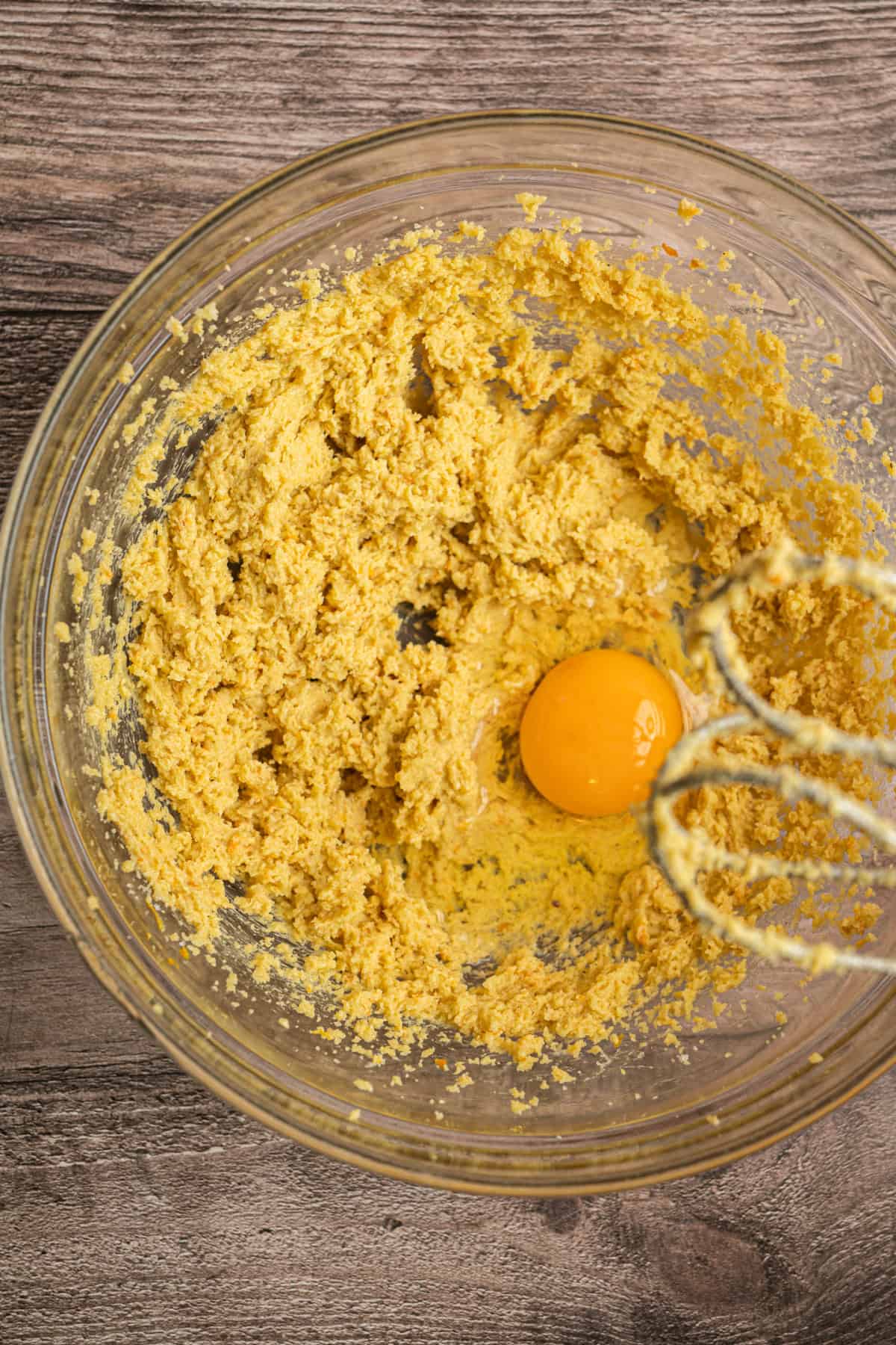 A glass mixing bowl containing yellow orange chocolate cookies dough and a whole egg, with a hand mixer partially visible on the side, sits on a wooden surface.
