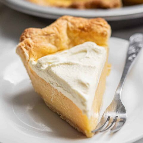 A slice of old-fashioned lemon cream pie with a flaky crust is served on a white plate. The pie is topped with a generous layer of whipped cream, and a fork rests beside it.