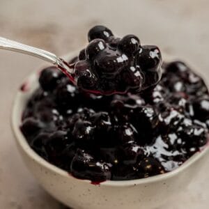 A spoonful of glossy, dark blueberry compote—perfect as a blueberry cake filling—is held above a bowl filled with the same compote, set against a neutral background.