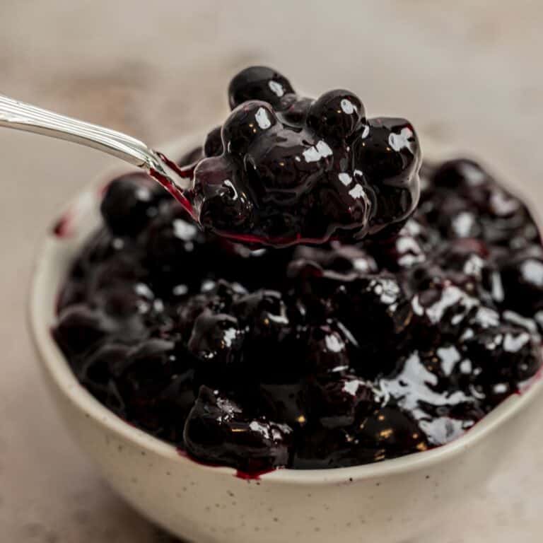 A spoonful of glossy, dark blueberry compote—perfect as a blueberry cake filling—is held above a bowl filled with the same compote, set against a neutral background.