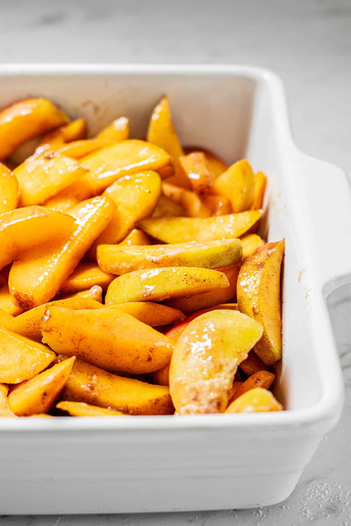 Sliced peaches coated in cinnamon and sugar are arranged in a white baking dish, ready to be baked. The dish sits on a light-colored countertop.