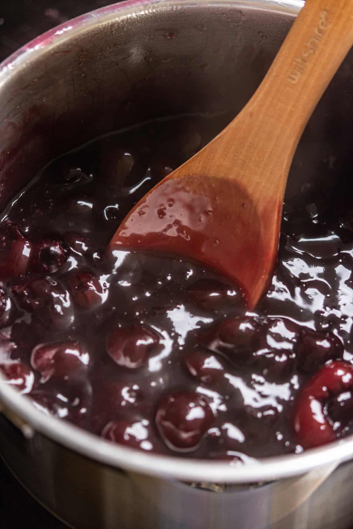 A close-up of a saucepan filled with glossy cherry pie filling, perfect for swirling into homemade black forest ice cream, being stirred with a wooden spoon.