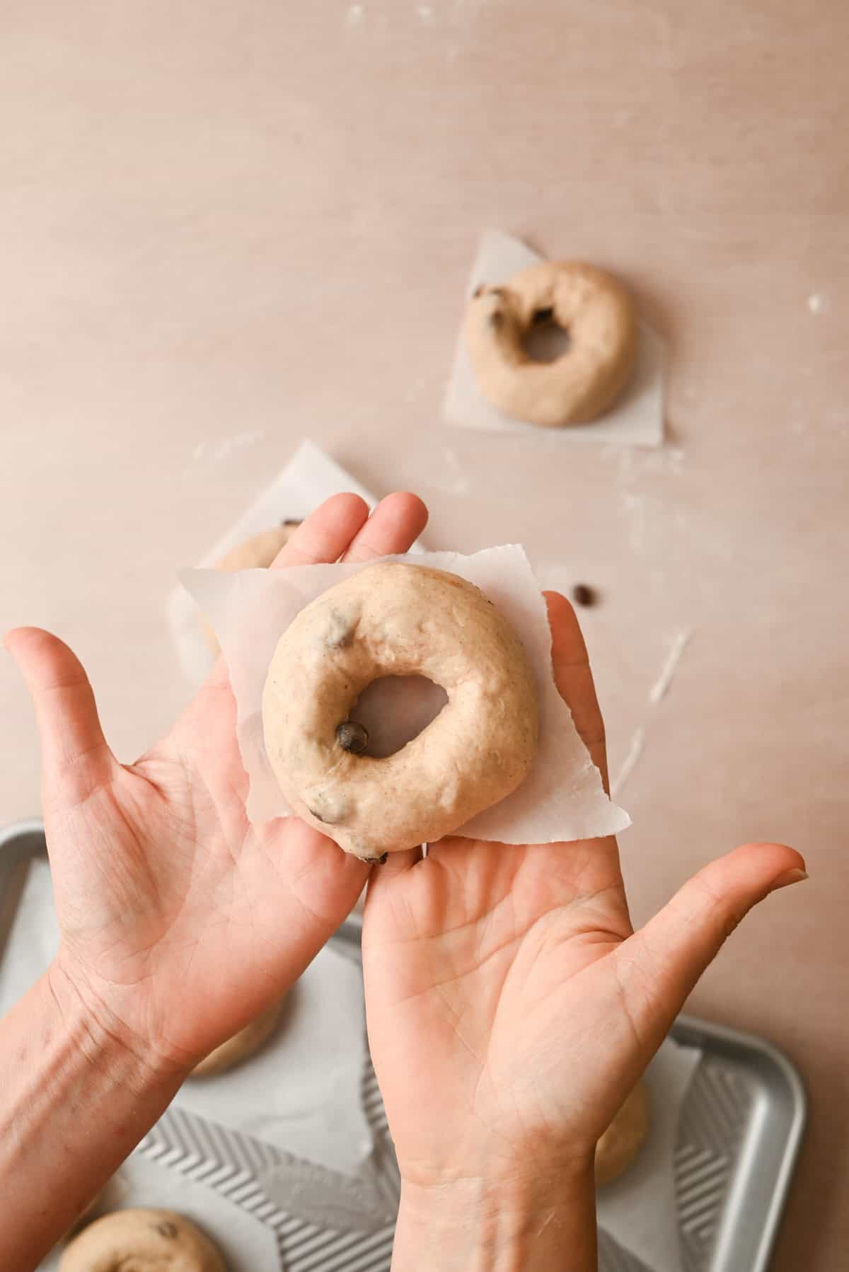 Two hands hold a raw cinnamon raisin bagel on a small piece of parchment paper above a baking tray, with more unbaked bagels on pieces of parchment in the background.