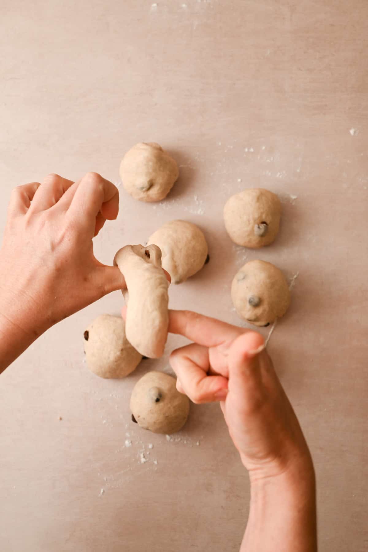 Two hands are shaping a piece of dough with chocolate chips, while several cinnamon raisin bagels and other dough balls rest on a floured surface in the background.