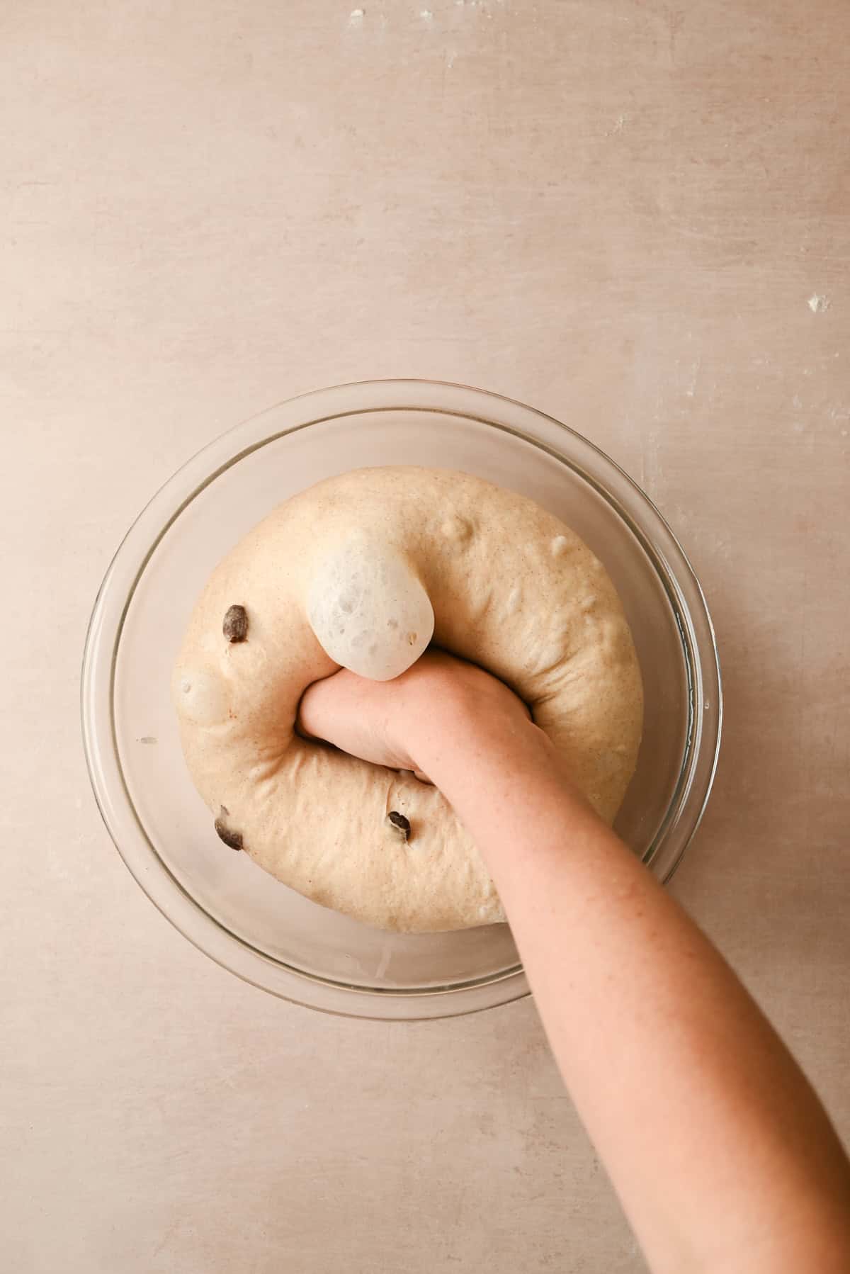A hand punches down risen chocolate chip bread dough in a glass bowl on a light beige surface, perfect for making treats like cinnamon raisin bagels.