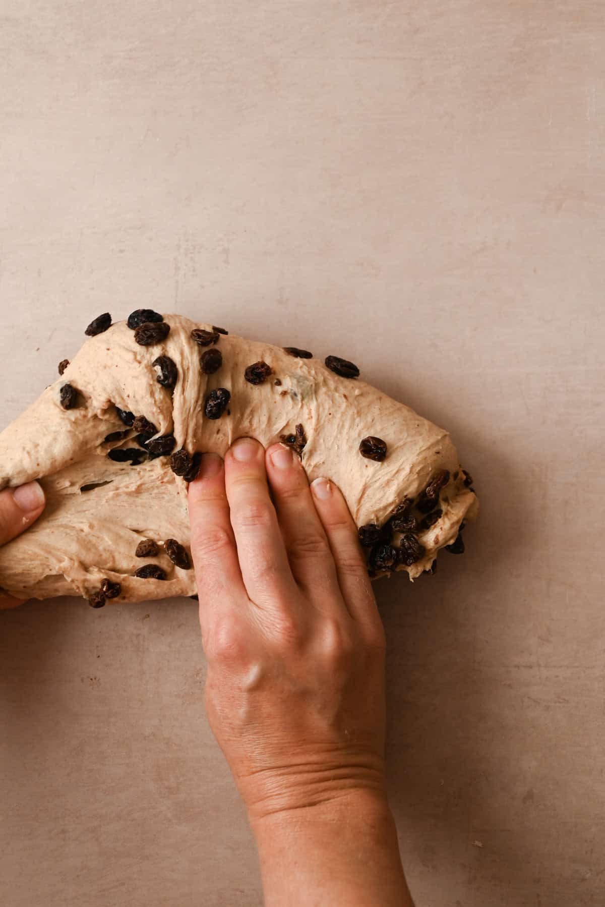 Hands kneading bread dough with raisins on a light brown surface, preparing it for delicious cinnamon raisin bagels.