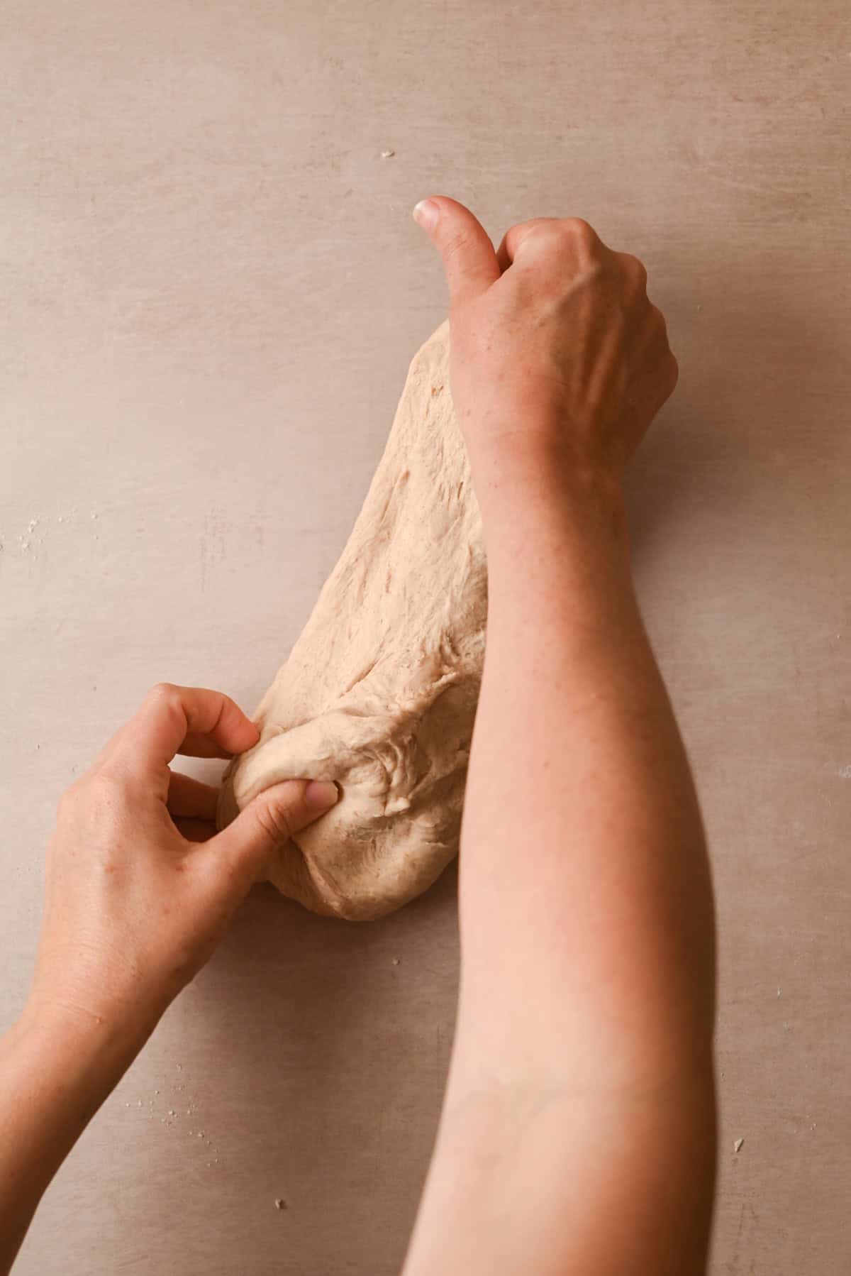 Two hands stretch and fold a piece of cinnamon raisin bagel dough on a light-colored surface, showing the process of kneading and shaping.