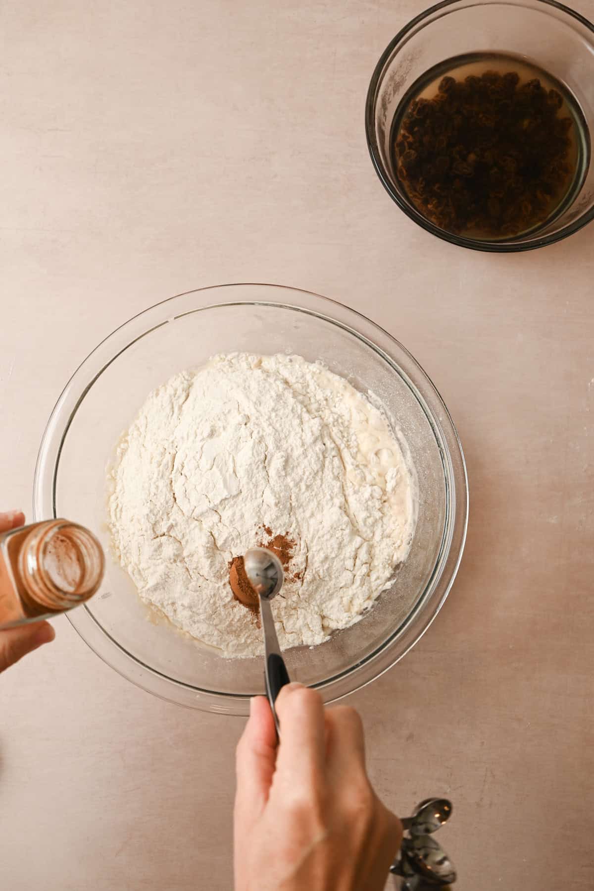 A hand adds ground cinnamon from a jar to a bowl of flour with a measuring spoon, prepping dough for cinnamon raisin bagels. Another glass bowl with soaked raisins sits nearby on a light countertop.