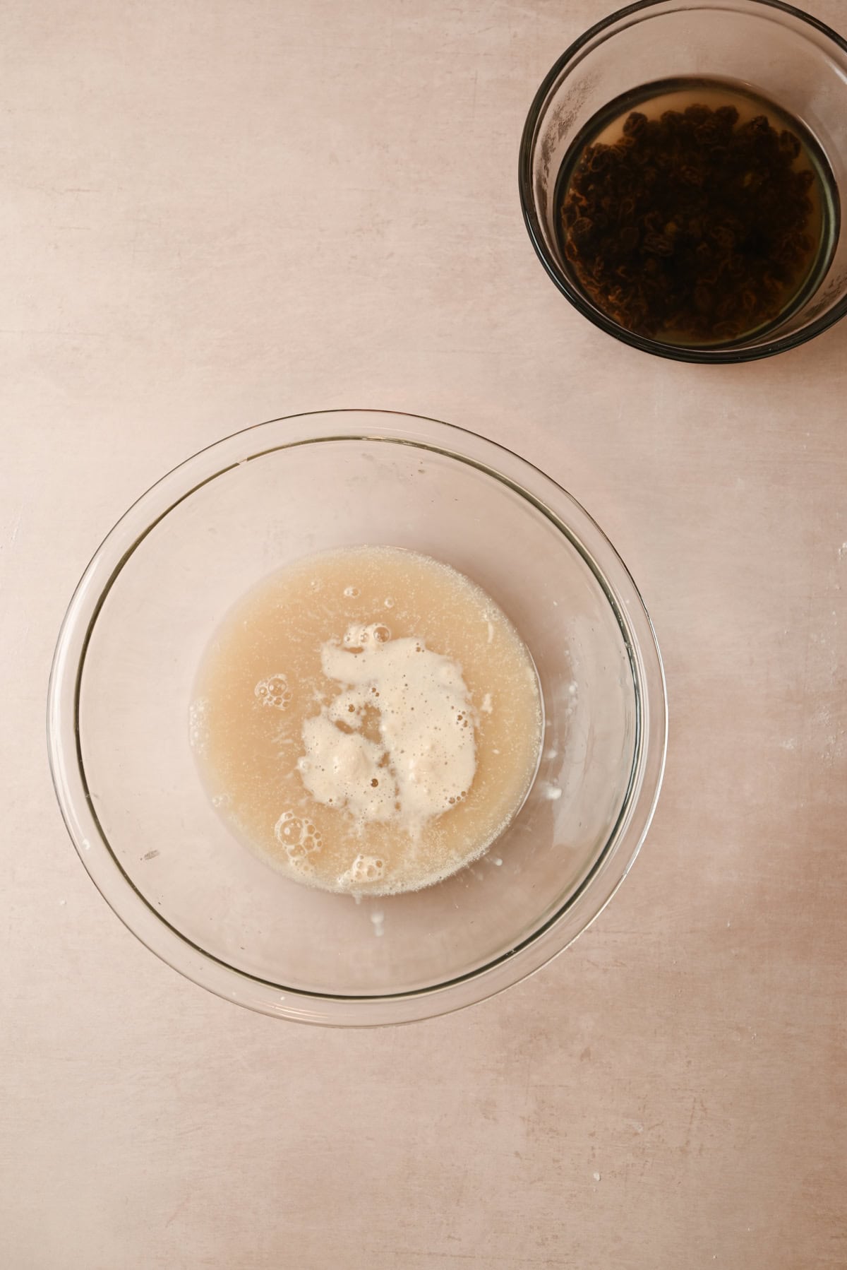 Two glass bowls on a light surface: the larger one holds a bubbling yeast mixture, while above it, the smaller bowl soaks raisins—both essential steps in making aromatic cinnamon raisin bagels.