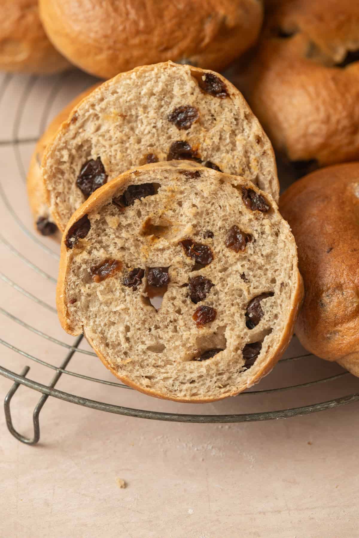 A close-up of a sliced cinnamon raisin bagel on a cooling rack, showing its airy texture and raisins inside. Other whole buns are visible in the background.