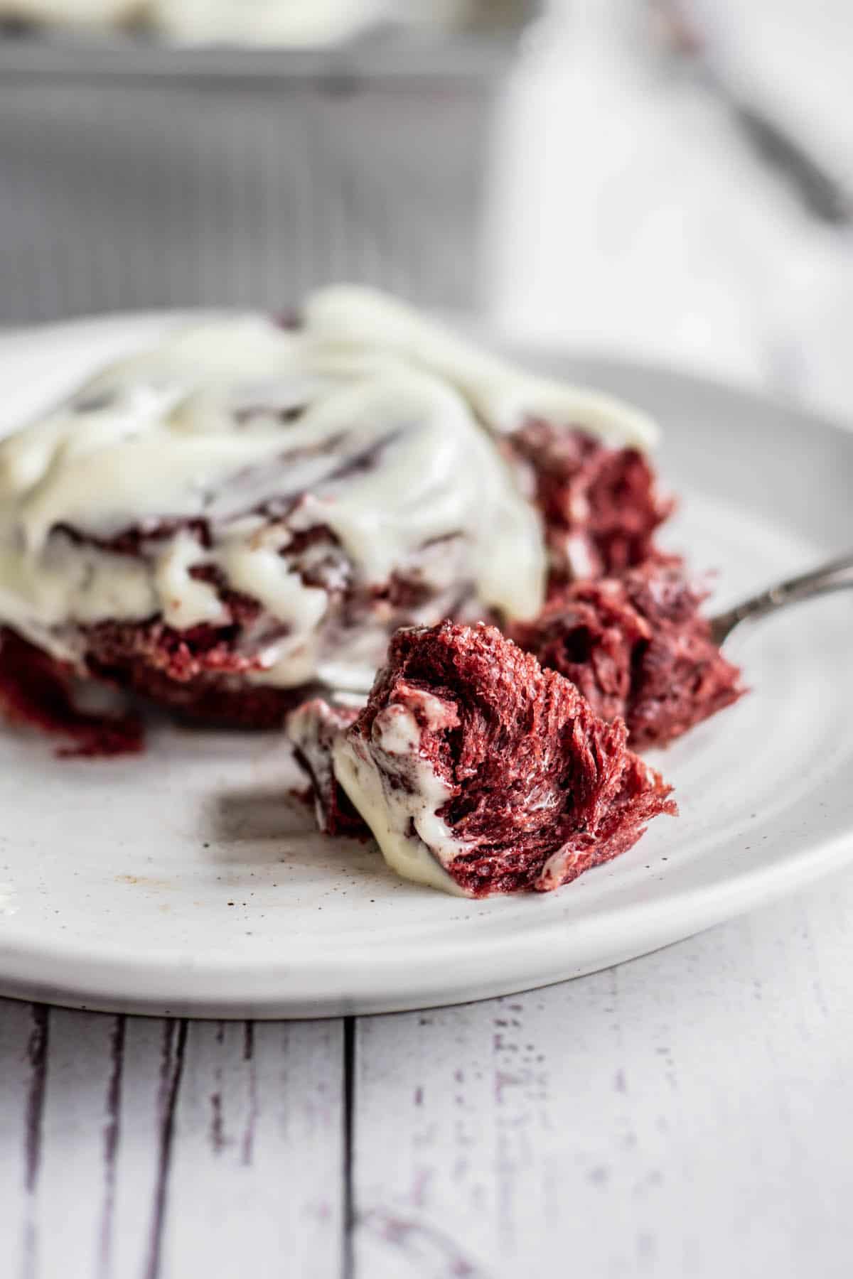 a bitten red velvet cinnamon roll with cream cheese frosting on a plate with a fork.