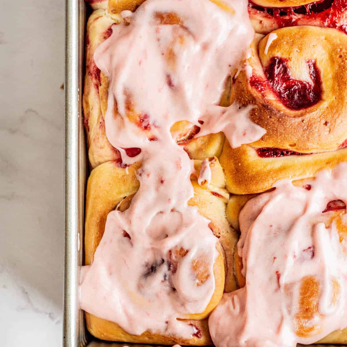 Top-down picture of strawberry cinnamon rolls with cream cheese icing spread over in a baking pan.