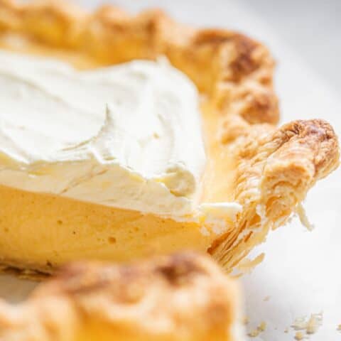 Close-up of a creamy pie with a golden, flaky crust like you'd find in the best Flaky Pie Crust Recipe. A slice has been cut, revealing a thick, smooth filling topped with whipped cream. The light and crispy crust perfectly complements the rich, pale yellow interior.