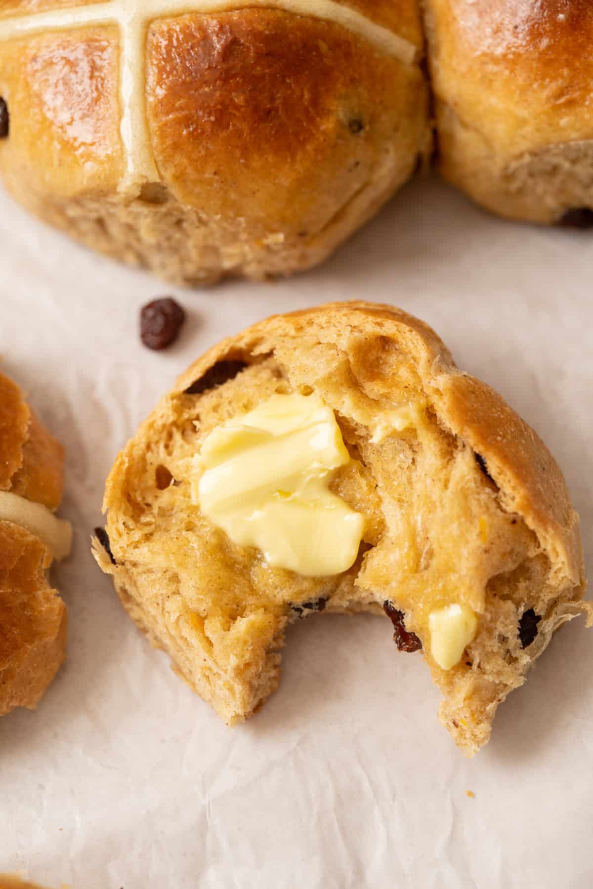 A close-up of a partially eaten Brioche Hot Cross Bun with a pat of melting butter on top, showing a soft, fluffy interior with visible raisins. Other whole buns are visible in the background on parchment paper.