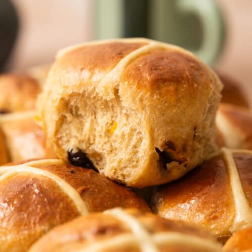 A close-up of freshly baked Brioche Hot Cross Buns, with one bun on top showing a bite taken out. The buns are golden brown with white icing crosses, and a green mug is blurred in the background.