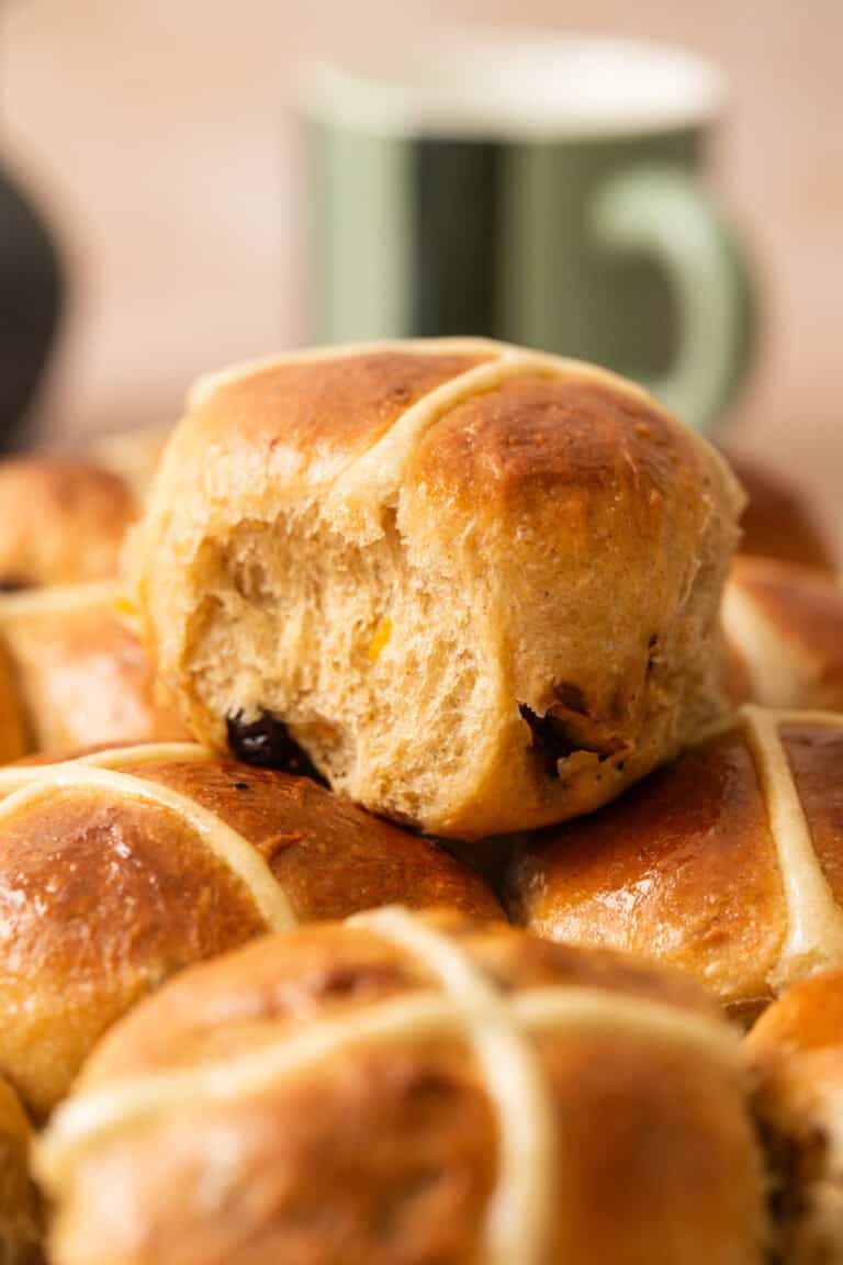 A close-up of freshly baked Brioche Hot Cross Buns, with one bun on top showing a bite taken out. The buns are golden brown with white icing crosses, and a green mug is blurred in the background.