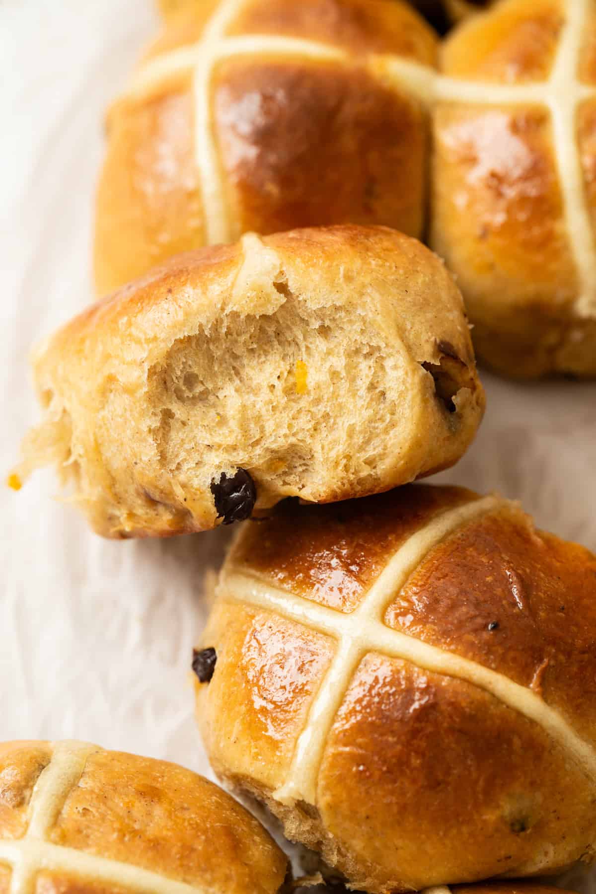 A close-up of golden-brown Brioche Hot Cross Buns, one with a bite taken out, showing a soft interior with visible raisins and a glossy finish, decorated with white icing crosses on top.