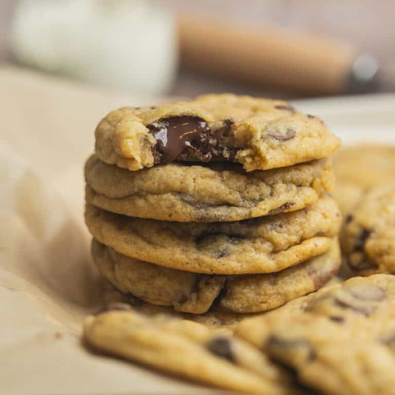 A stack of five chewy sourdough discard chocolate chip cookies sits on parchment paper, the top cookie missing a bite to reveal gooey melted chocolate chips inside. More cookies are scattered in the background.