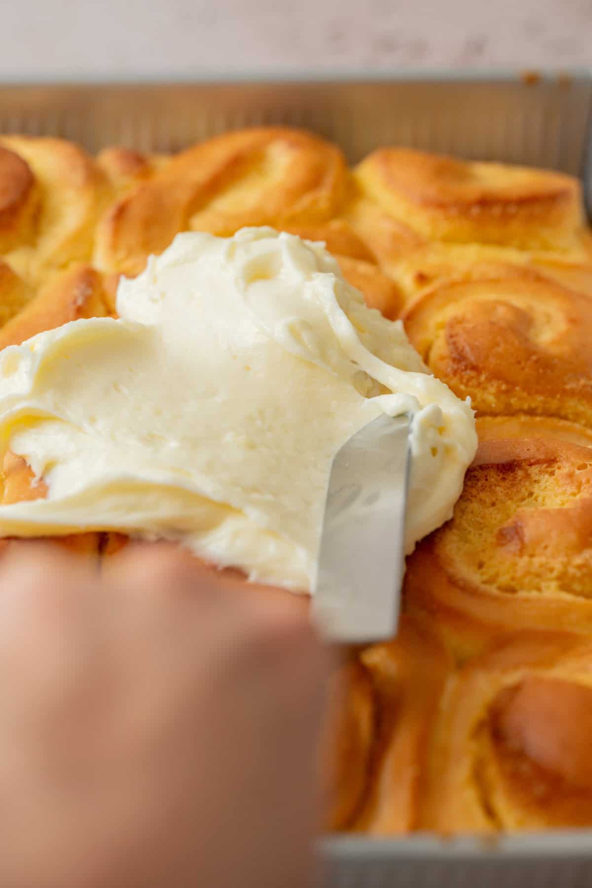 A hand spreads creamy white frosting over a tray of golden brown almond sweet rolls using a metal spatula.