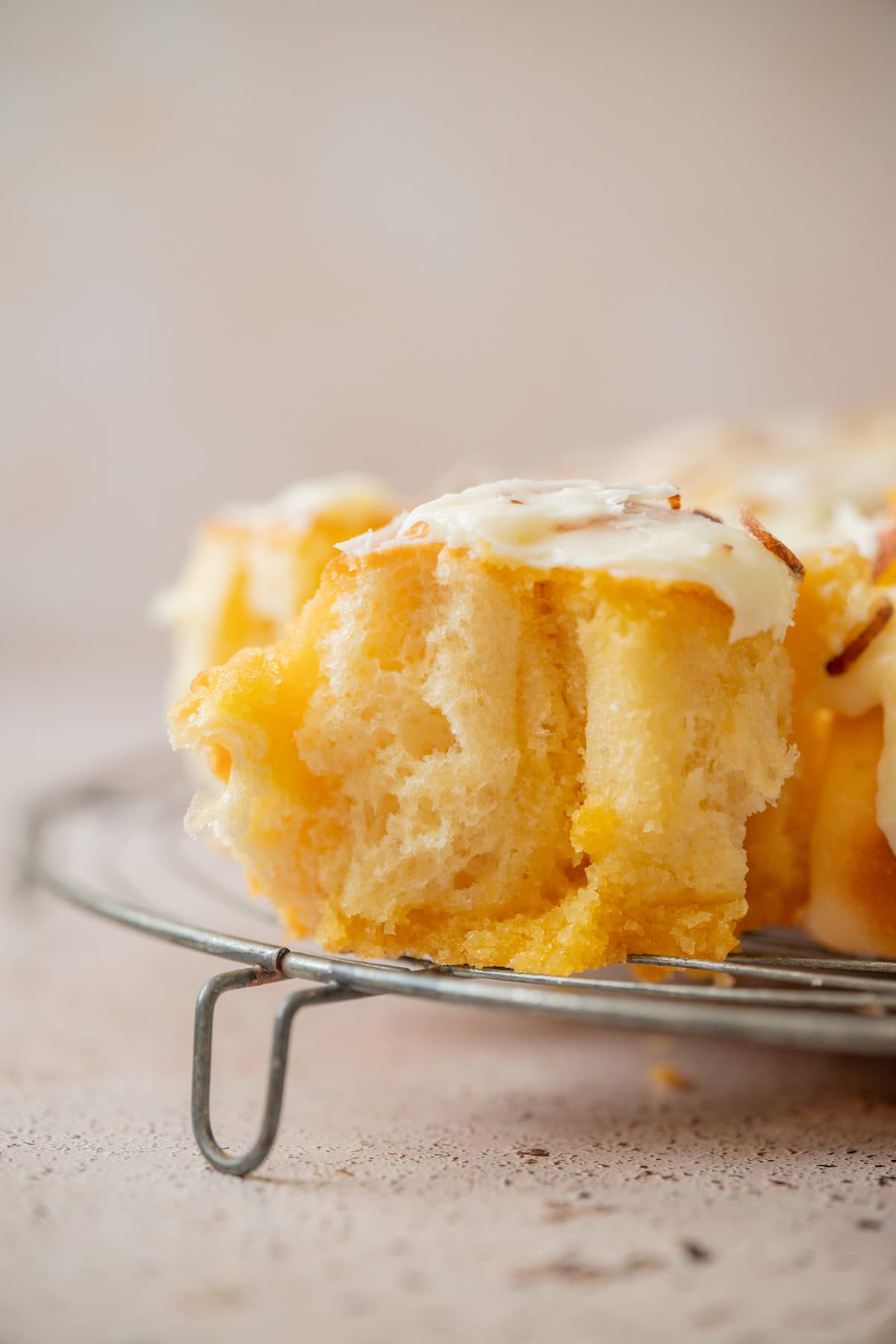 A close-up of a fluffy, golden almond sweet roll topped with creamy white icing, sitting on a metal cooling rack with a soft, neutral background.