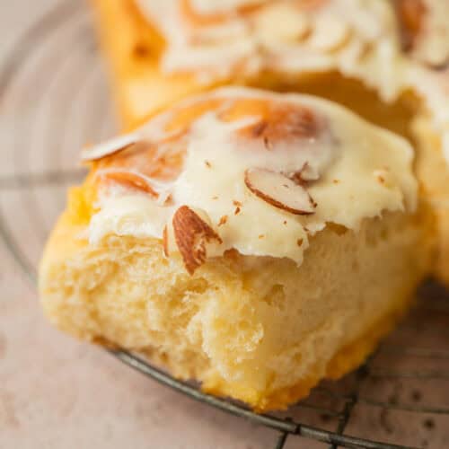 A close-up of a slice of frosted bread topped with sliced almonds, resting on a metal cooling rack. The creamy frosting and fluffy texture evoke the irresistible appeal of almond sweet rolls.