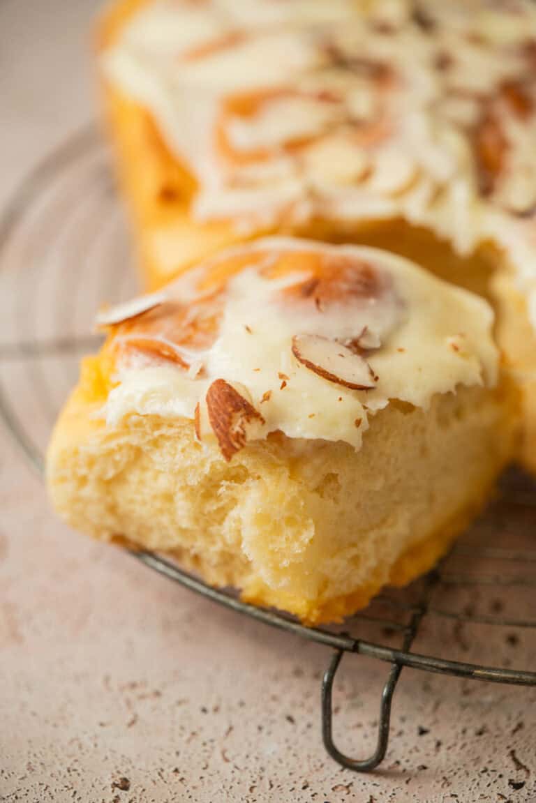 A close-up of a slice of frosted bread topped with sliced almonds, resting on a metal cooling rack. The creamy frosting and fluffy texture evoke the irresistible appeal of almond sweet rolls.