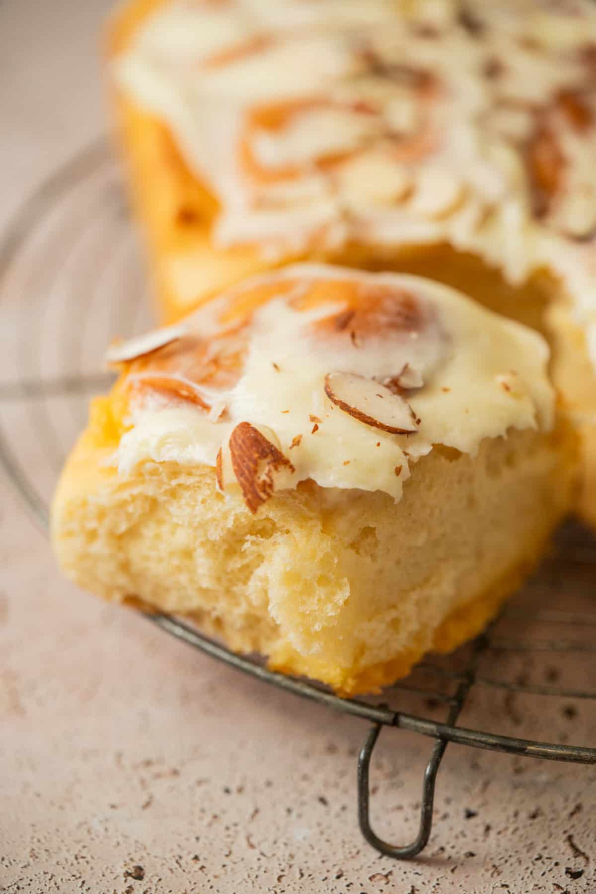 A close-up of a slice of frosted bread topped with sliced almonds, resting on a metal cooling rack. The creamy frosting and fluffy texture evoke the irresistible appeal of almond sweet rolls.