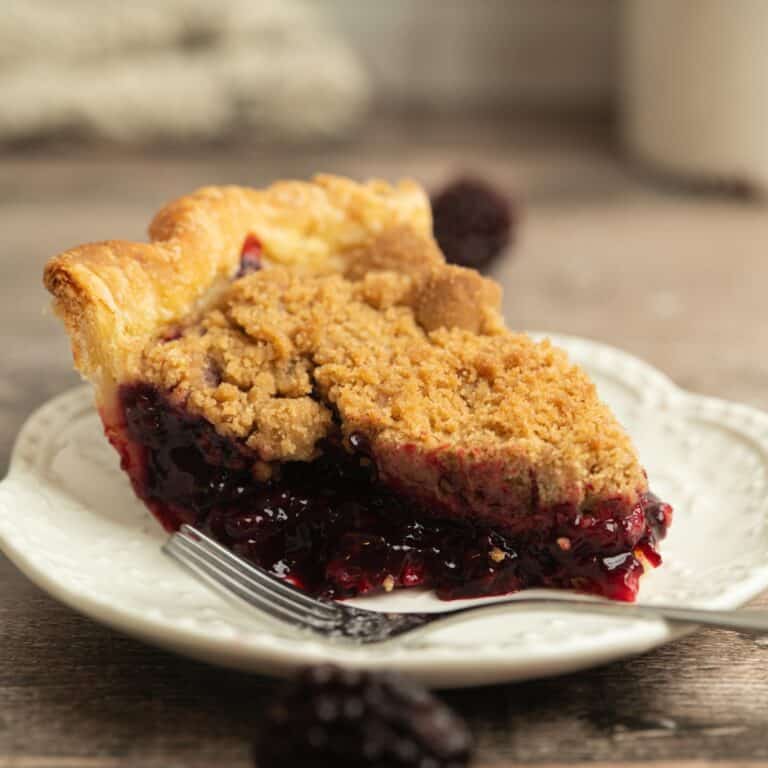 A slice of Blackberry Crumble Pie sits on a white plate with a fork. The golden, crumbly topping covers a thick, juicy blackberry filling, while whole blackberries in the background complement this tempting dessert.