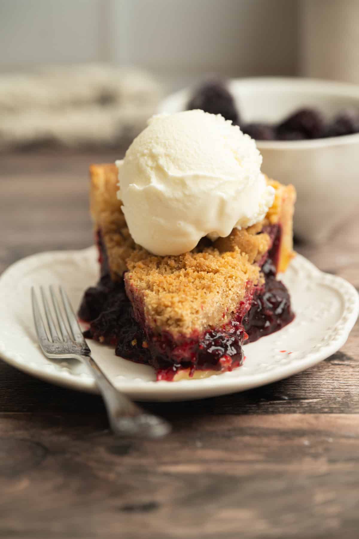 A slice of Blackberry Crumble Pie topped with a scoop of vanilla ice cream sits on a white plate with a fork, with a bowl of berries in the background on a wooden table.