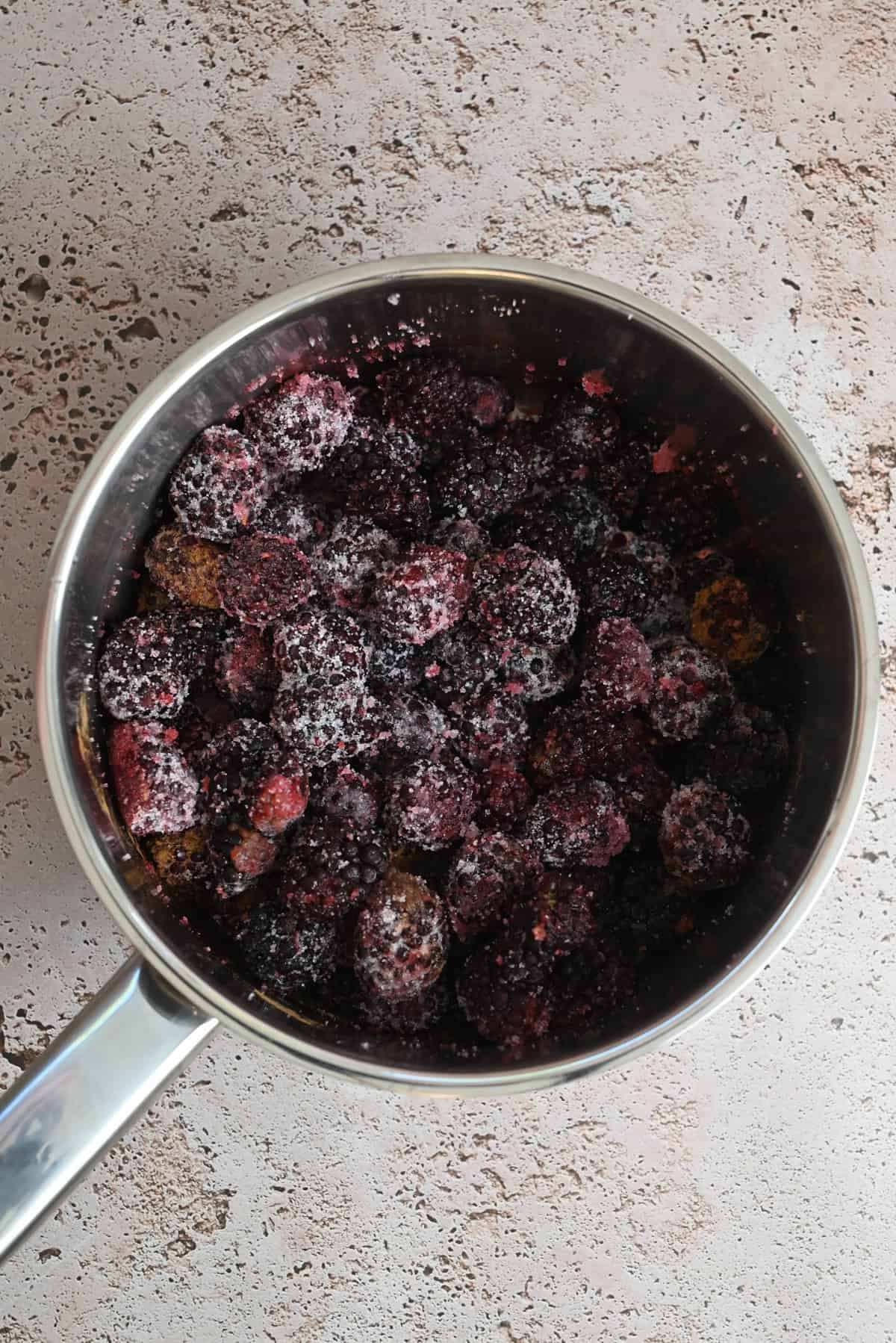 A stainless steel saucepan filled with frozen mixed berries and granulated sugar, perfect for making Blackberry Crumble Pie, sits on a light textured surface.