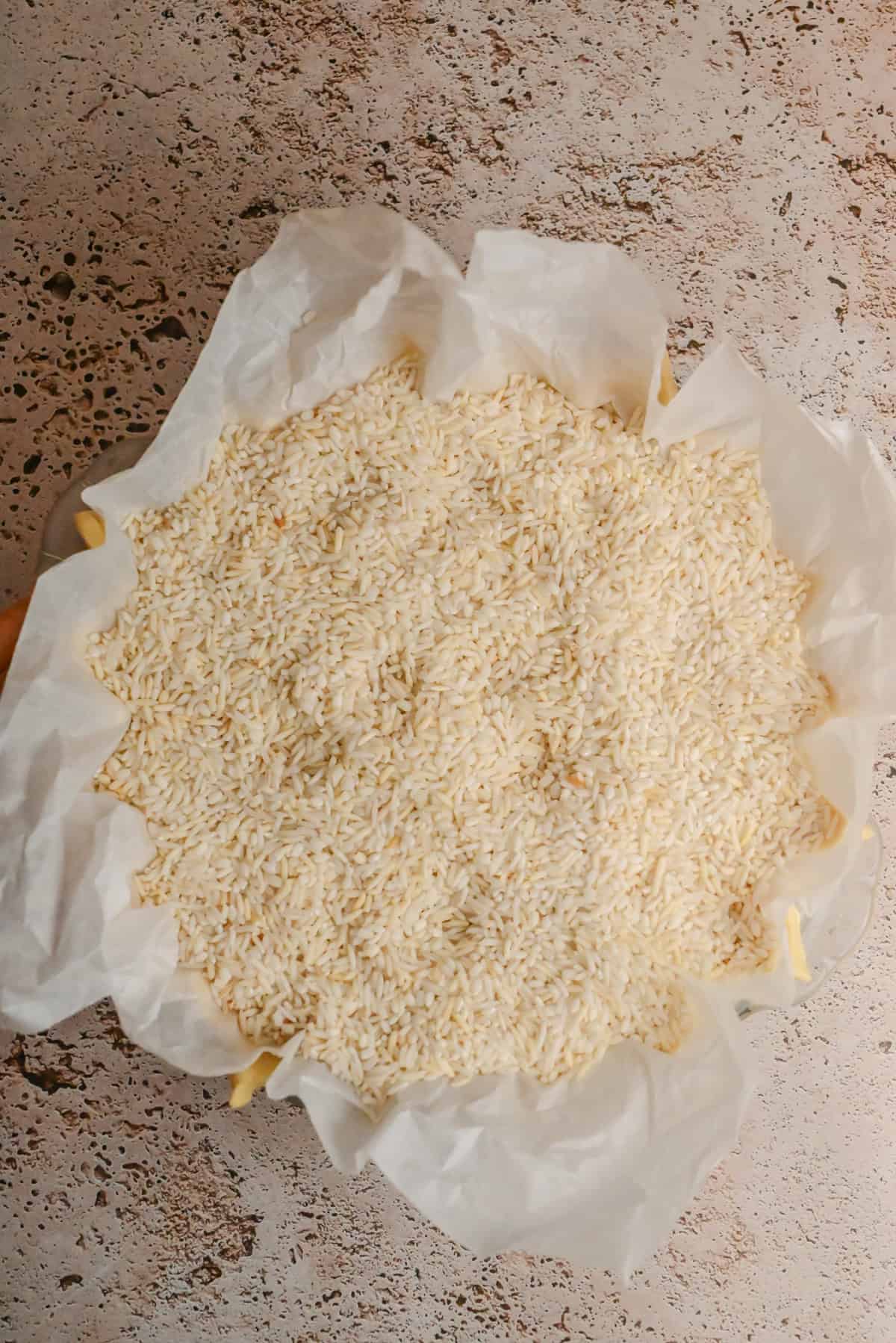 A round baking dish lined with parchment paper, prepped for Blackberry Crumble Pie, is filled with uncooked white rice and sits on a lightly speckled countertop.