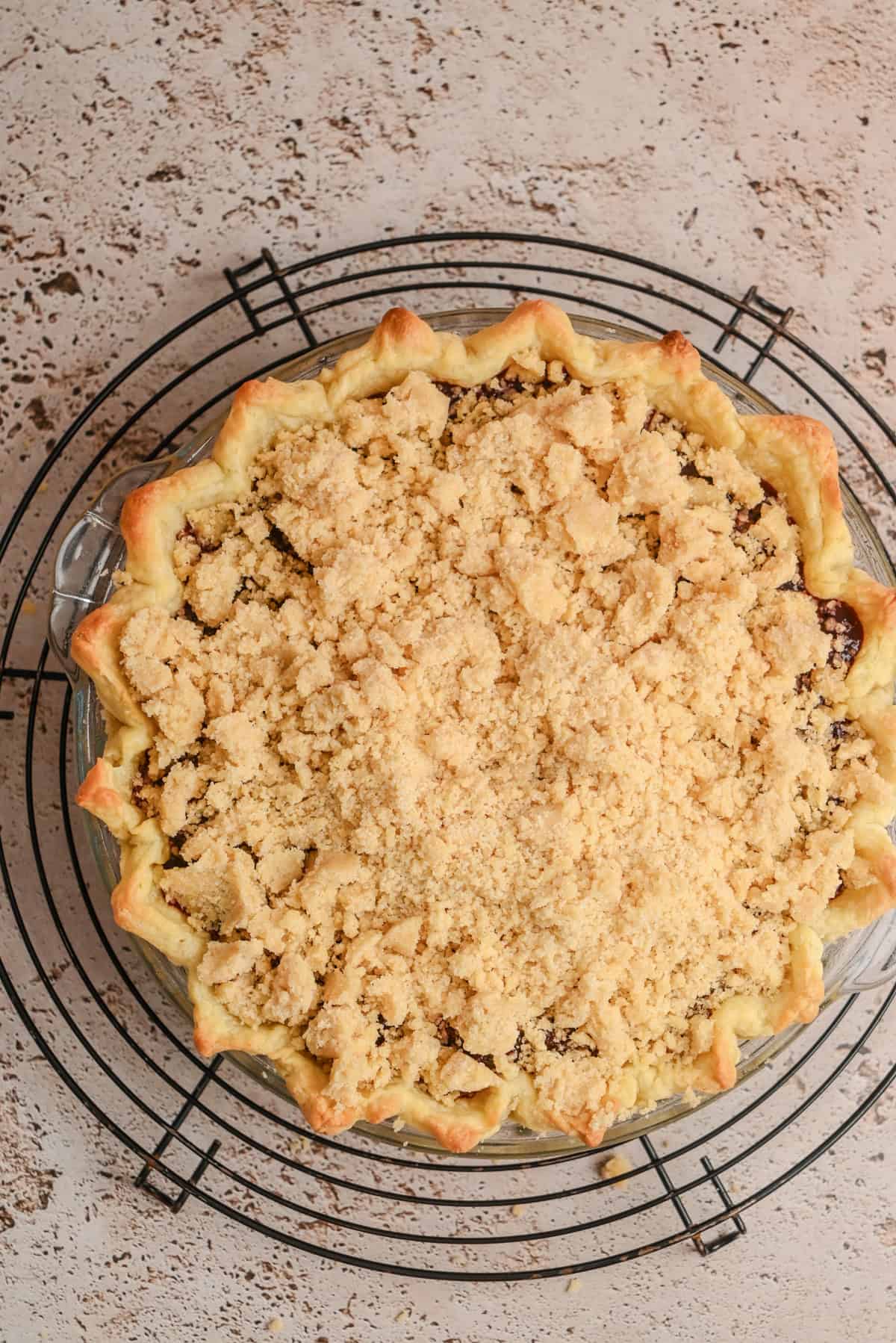 A freshly baked Blackberry Crumble Pie sits on a round wire cooling rack, its golden, crimped edge and generous layer of streusel topping showcased on a speckled beige countertop.