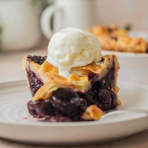 A slice of cherry pie with frozen cherries features a golden crust and dark cherry filling, sitting on a plate and topped with a scoop of vanilla ice cream.