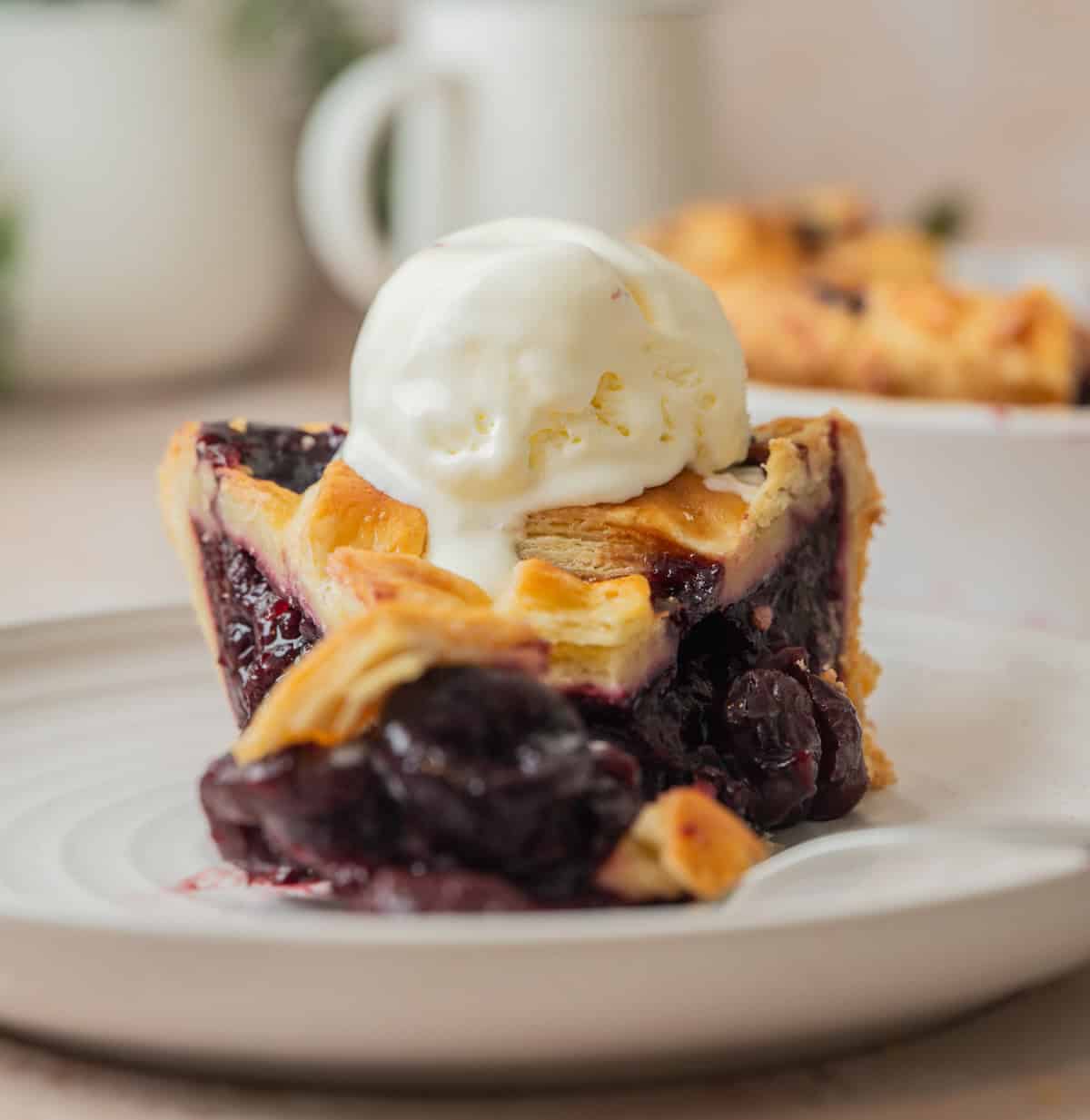 A slice of cherry pie with frozen cherries features a golden crust and dark cherry filling, sitting on a plate and topped with a scoop of vanilla ice cream.