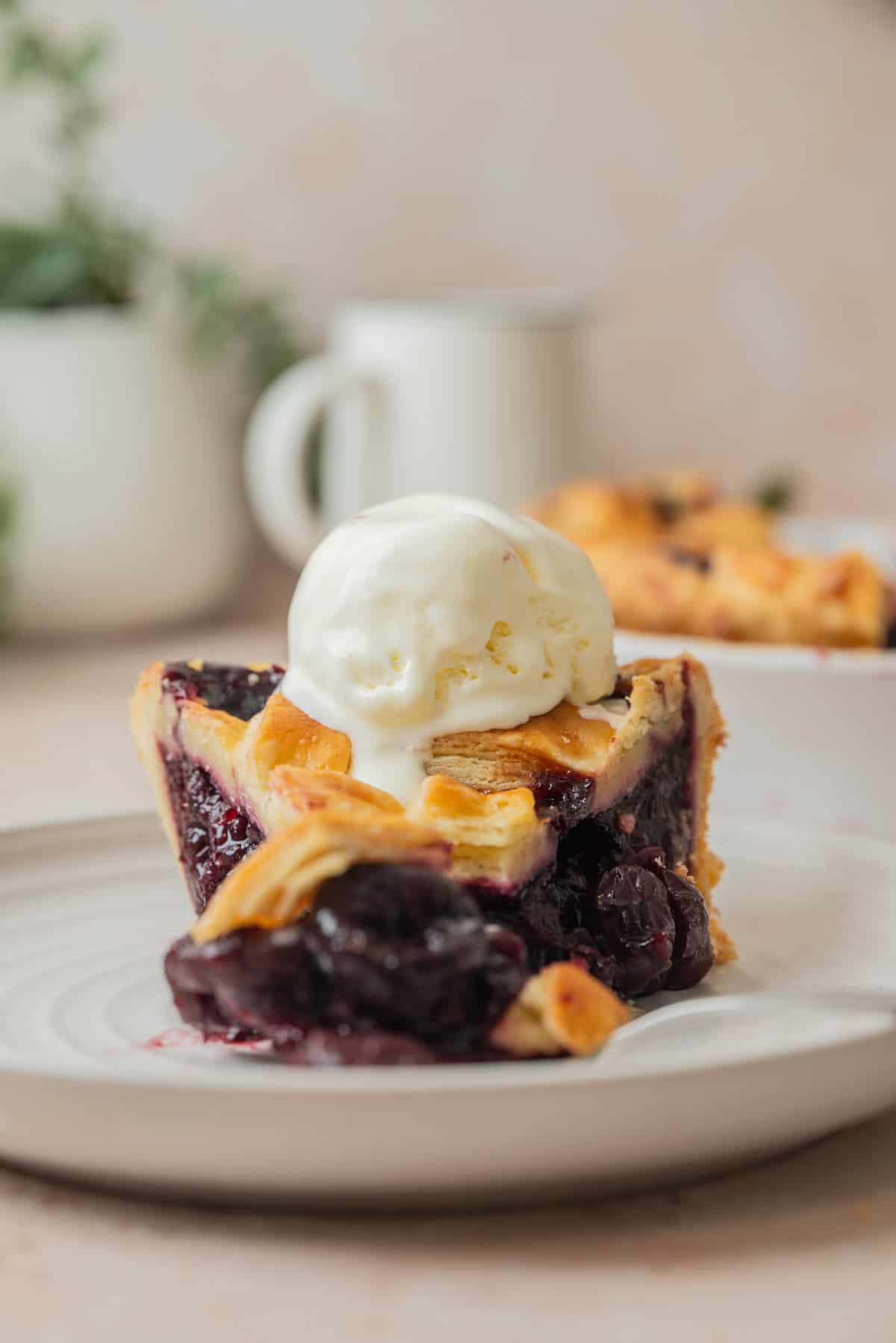 A slice of cherry pie with frozen cherries, topped with a scoop of vanilla ice cream, sits on a white plate. In the background, a blurred mug and another slice add to the cozy scene.